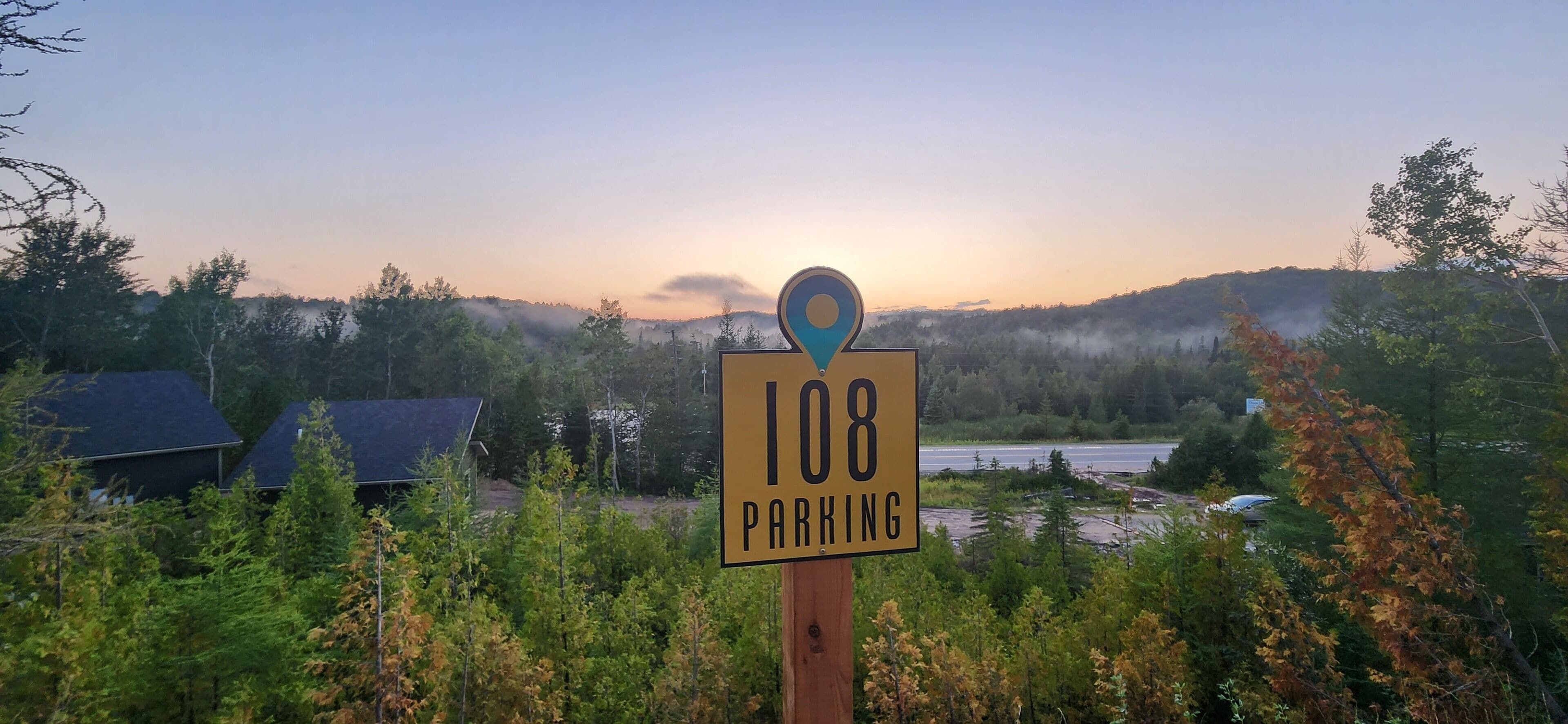 A parking sign with a location marker, surrounded by trees and a sunset in the background.