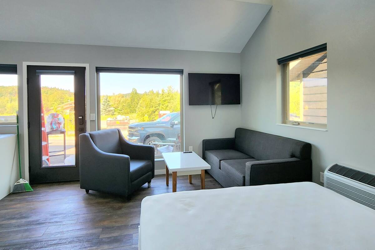 Quarto de hotel moderno com uma cama grande, um sofá, uma mesa pequena e uma TV. Janelas amplas oferecem vista para o exterior.