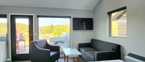 A modern hotel room with a large bed, a sofa, a small table, and a TV. There are large windows offering a view of the outdoors.