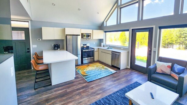 Una cocina moderna con una isla central, un área de comedor con sofá y ventanas amplias con vistas al exterior.