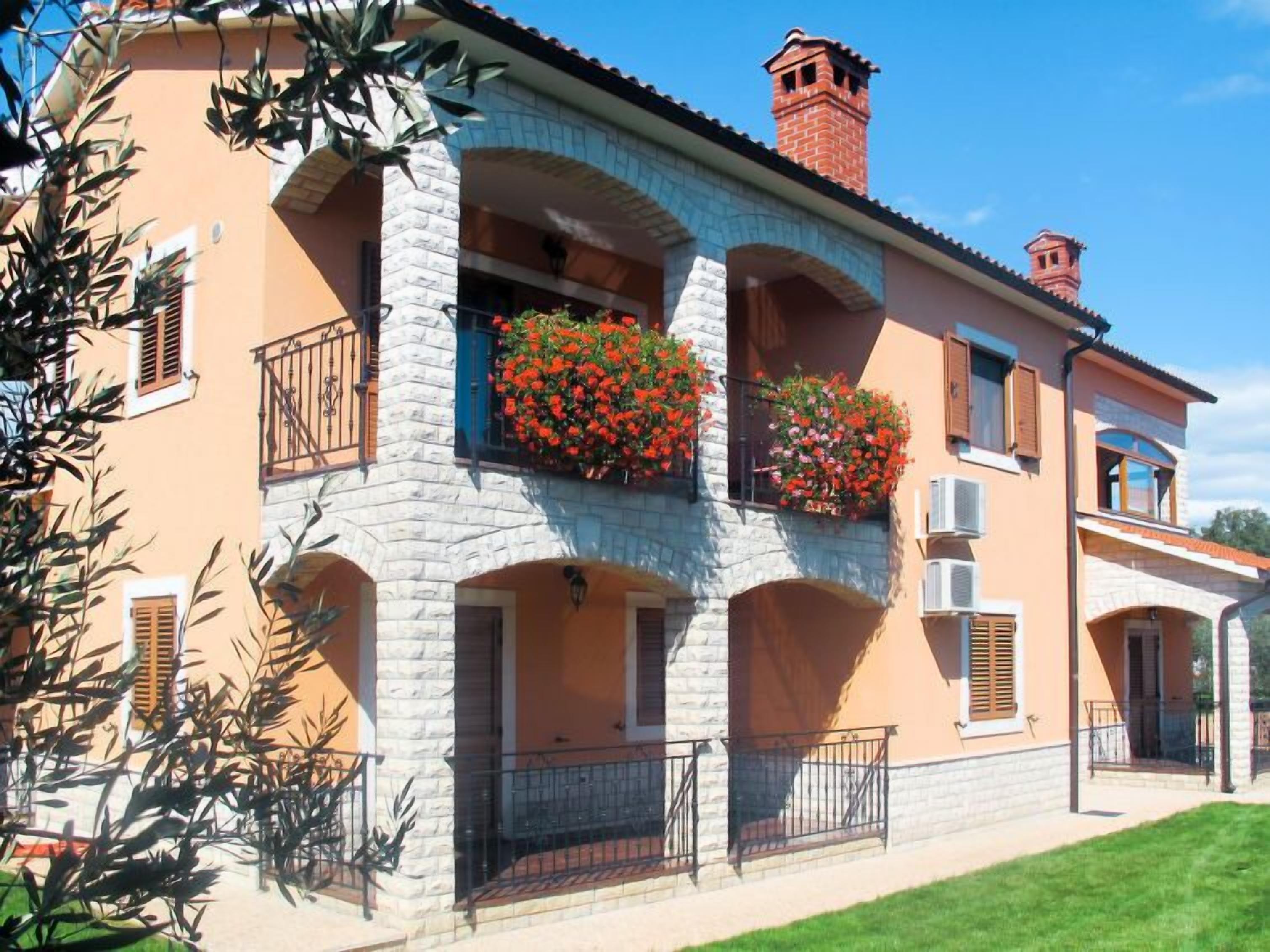 Un edificio de dos pisos con fachada de piedra y estuco, balcones con macetas de flores y un cielo despejado.