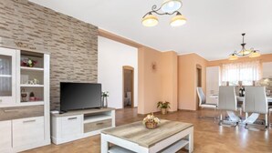 Un salon moderne doté d’un téléviseur à écran plat, d’une table basse en bois et d’un coin repas avec des chaises blanches et un lustre.