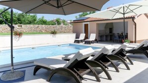 Une piscine entourée de chaises longues et de parasols, le tout délimité par un mur de pierre et bordé d’un bâtiment dont le toit est recouvert de tuiles.