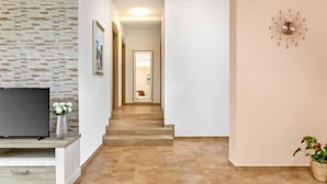 Un salon moderne avec un téléviseur à écran plat posé sur un meuble en bois, un mur à la texture pierre, un escalier menant à une autre pièce et une horloge au mur.