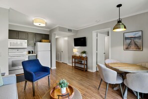 Eine moderne Küche mit weißen Geräten, eine Essecke mit einem Holztisch und -stühlen, und ein Wohnbereich mit einem blauen Sessel und einer Pflanze auf einem Tablett.