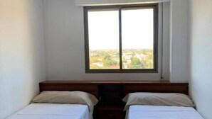 Quarto pequeno com duas camas de solteiro, ventilador de teto e janela com vista para a cidade.