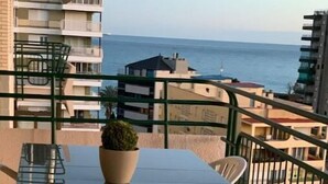 Uma varanda com mesa e cadeiras de vidro, um vaso de planta e vista para o mar.