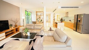 A modern living room with a sofa, coffee table, and TV, leading to a kitchen with a refrigerator and a dining area.