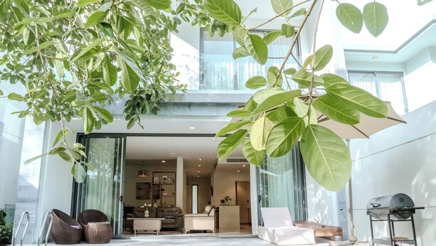 A modern outdoor pool area with a glass-enclosed living space, comfortable seating, and a barbecue setup.