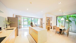 A modern kitchen with a central island, a dining area with a table and chairs, and large windows with a view of greenery.