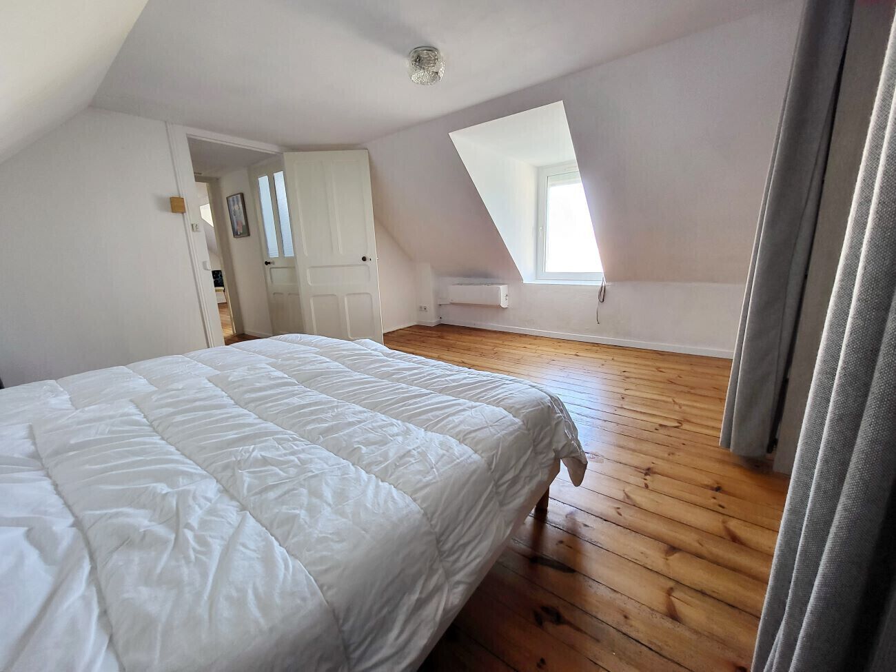 Une chambre avec un grand lit, un plancher en bois, une lucarne et une porte donnant sur une autre pièce.
