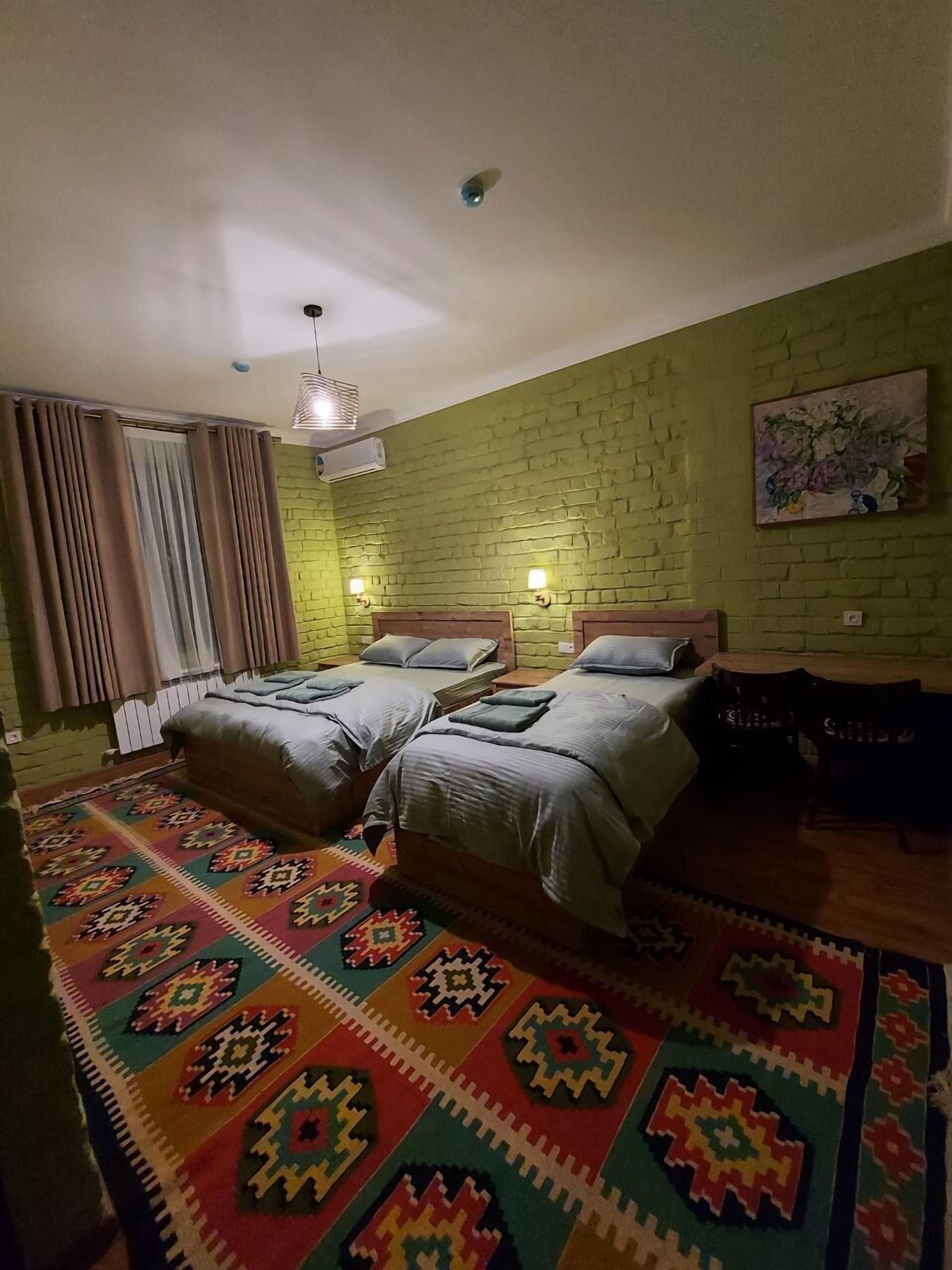 A room with two beds, a patterned rug, a green brick wall, and a painting.