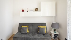 Ein modernes Wohnzimmer mit einem grauen Sofa, zwei gelben Kissen, einem weißen Beistelltisch mit einer Lampe und einer dekorativen Platte auf einem Regal darüber.