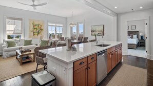 Una cocina moderna con una isla central, armarios de madera y lavavajillas. Hay una zona de comedor con mesa y sillas, y una sala de estar con sofá y mesita de centro.