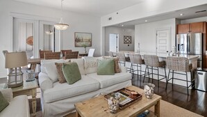 Una sala de estar moderna con un sofá, una mesa de centro y sillas de mimbre. Se ve, al fondo, una zona de cocina con un refrigerador y taburetes de bar.
