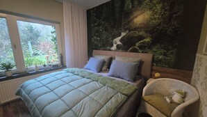 Une chambre avec un grand lit, une fenêtre avec des rideaux, une fresque murale représentant une scène de forêt et un fauteuil en osier avec un coussin.