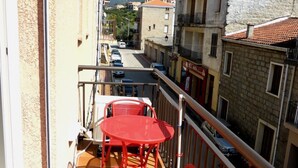 Ein Balkon mit einem roten Metalltisch und -stühlen, mit Blick auf eine Straße, an deren Seite Gebäude und ein Berg im Hintergrund zu sehen sind.