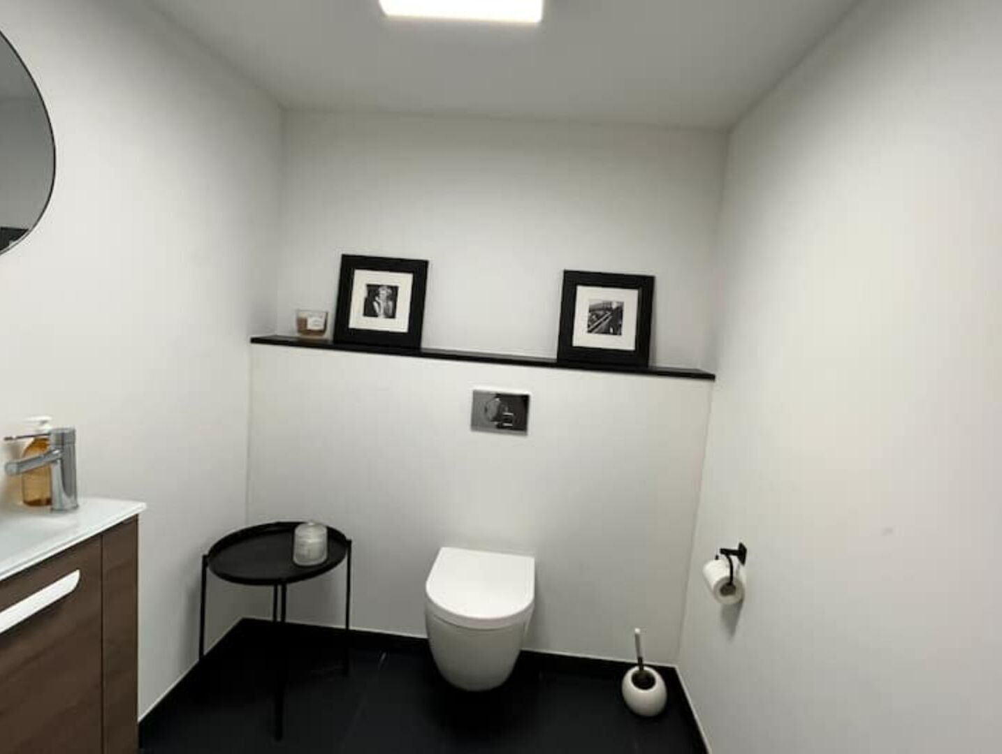 A small, modern bathroom with a white toilet, a black sink, and a small round table with a candle. There are two framed pictures on the wall above the toilet.