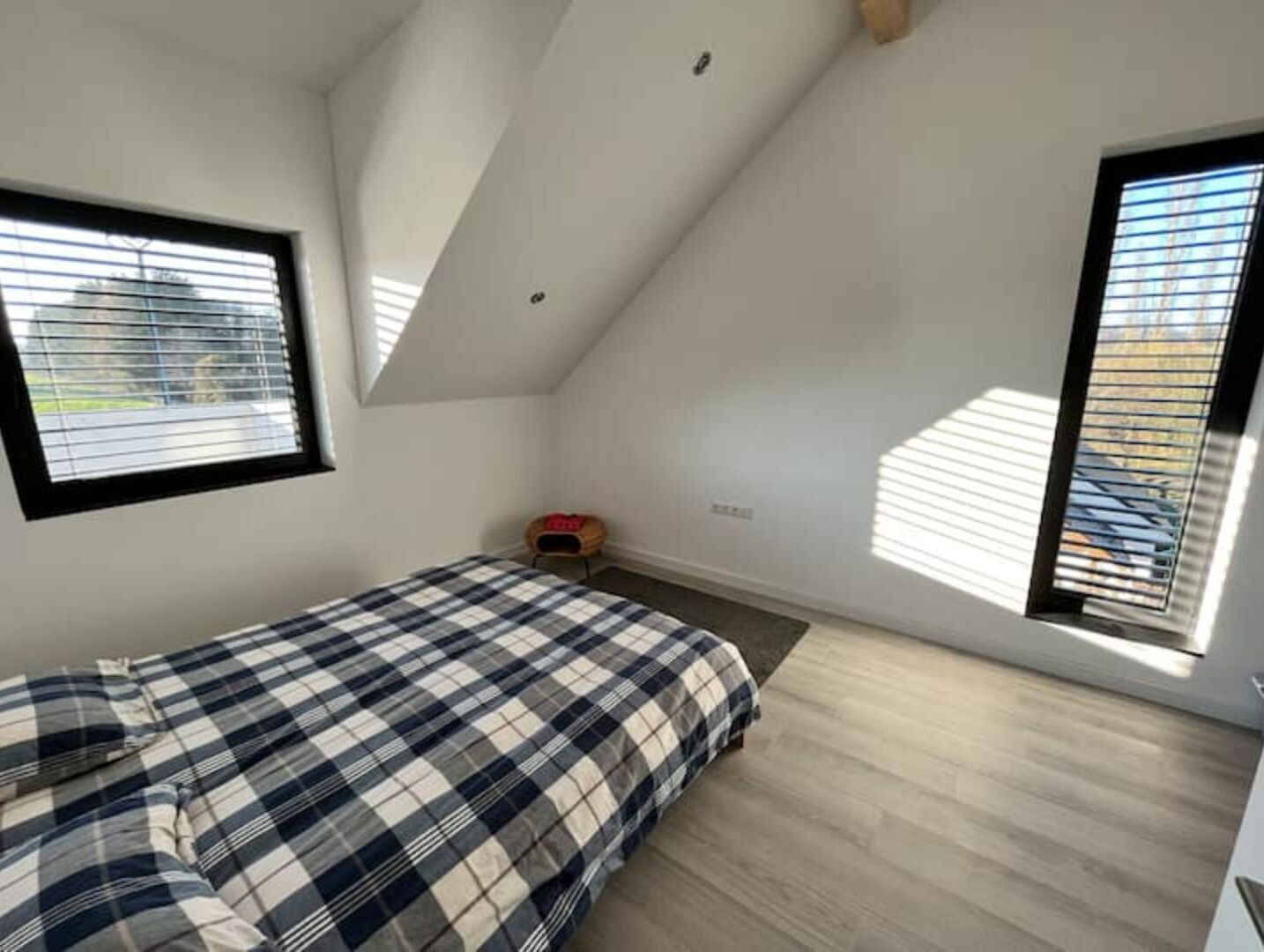 A small, well-lit room with a sloped ceiling, a bed with a plaid bedspread, a small wooden stool, and a window with blinds.