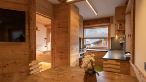 A wooden cabin interior with a wooden dining table, a kitchen area with a window, and a small potted plant on the table. - Casa Ca Dei Pini, surrounded by Nature (Cortina d´Ampezzo)