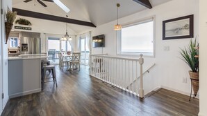 Une cuisine spacieuse dotée d’un îlot central, un coin repas avec une table et des chaises, un escalier avec une rampe blanche et une grande fenêtre qui laisse entrer la lumière naturelle.
