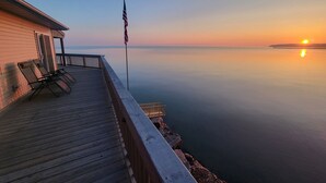 Une terrasse en bois avec deux chaises longues et un drapeau américain, donnant sur une étendue d’eau calme au coucher du soleil.