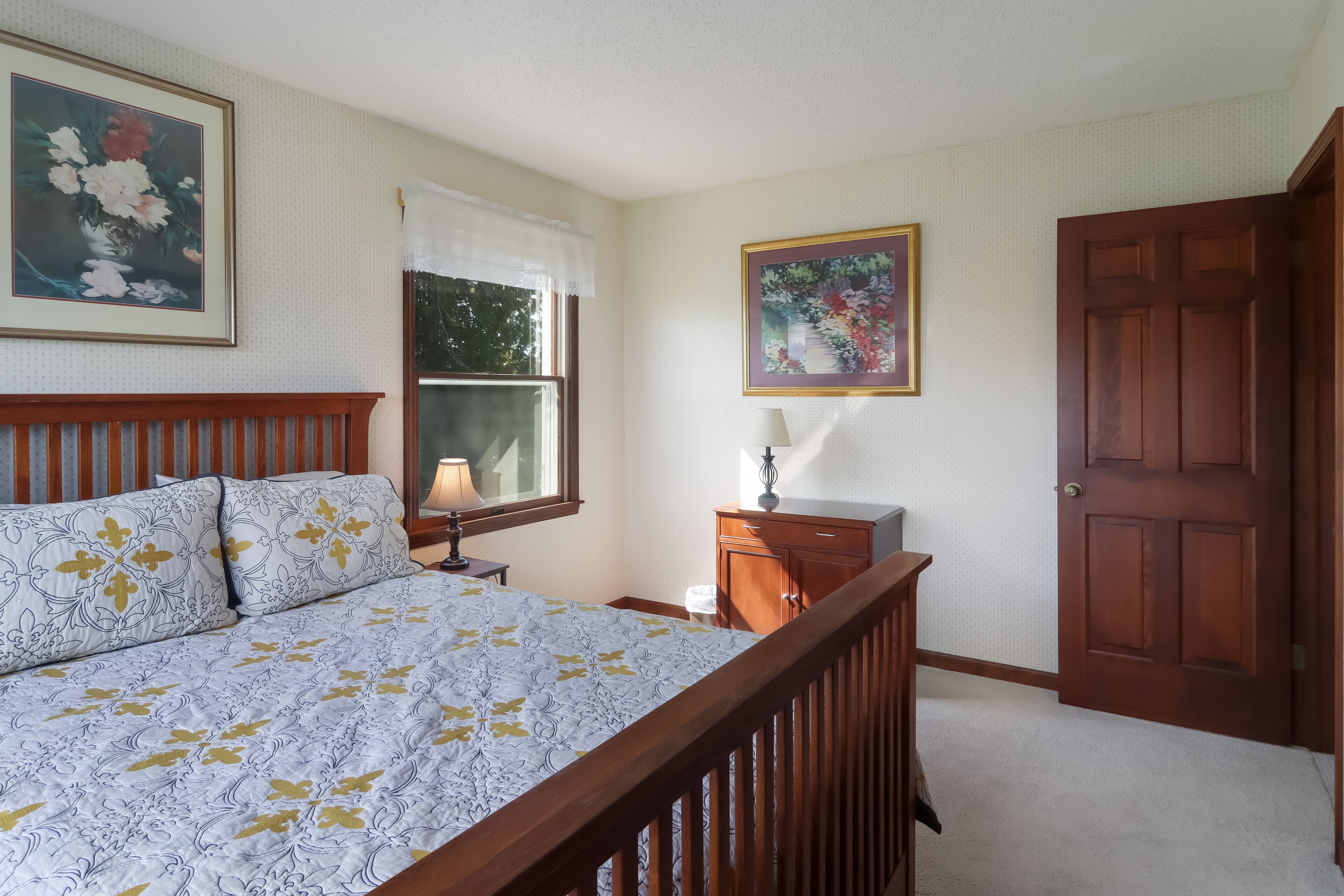 A bedroom with a bed, a nightstand, a dresser, a window with curtains, and a door.