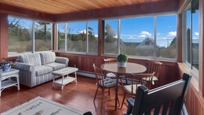 Un solárium con techo y paredes de madera, ventanas amplias, un sofá, una mesa de comedor con sillas y una planta pequeña en maceta.