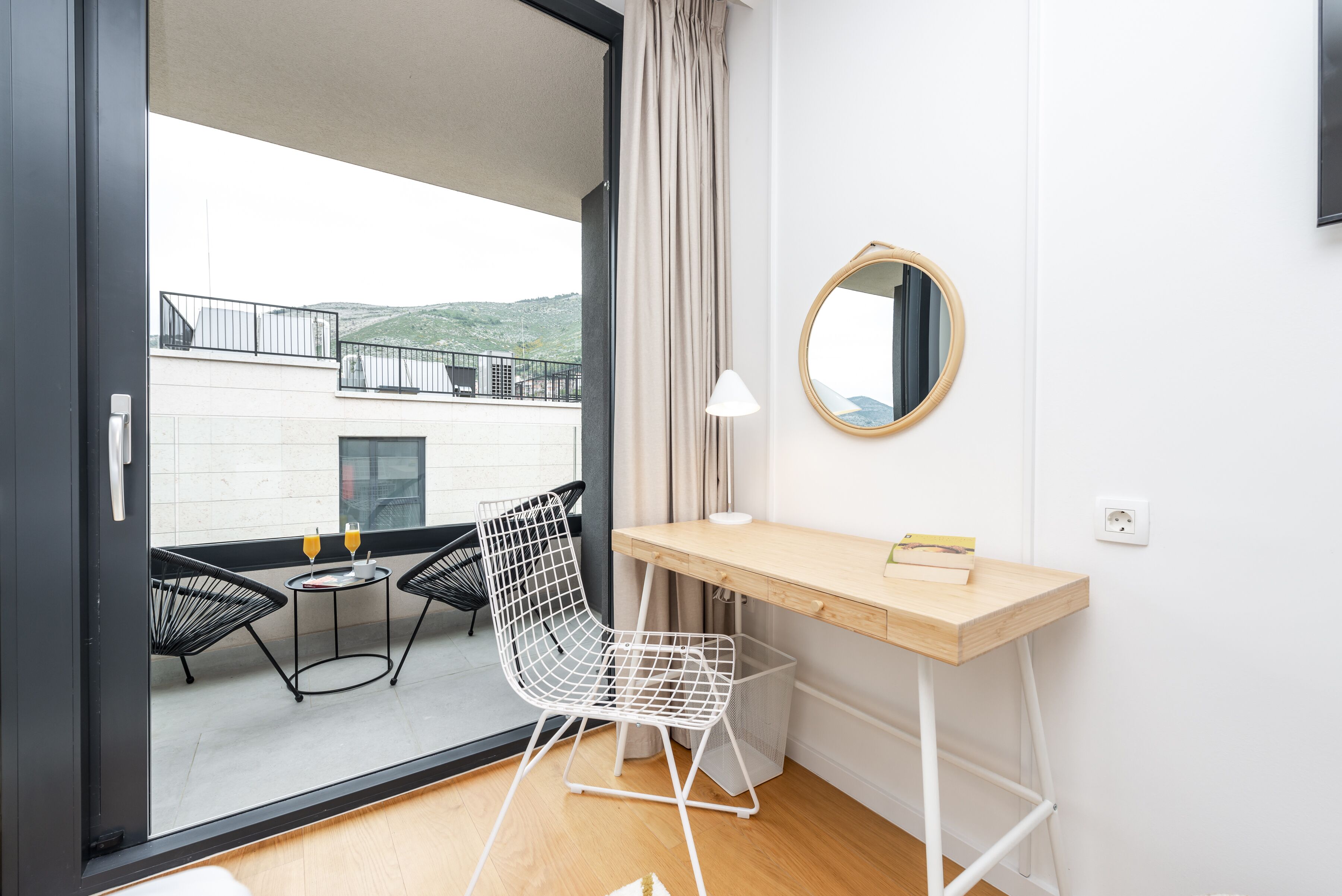 A modern room with a wooden desk, a white chair, a round mirror, and a view of a balcony with outdoor furniture.