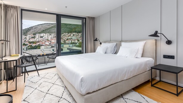 A modern hotel room with a large bed, a desk, and a view of a cityscape through floor-to-ceiling windows. - City Residences (Dubrovnik)
