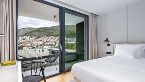 A hotel room with a bed, a desk, and a chair. There is a view of a city through a glass door. - City Residences (Dubrovnik)