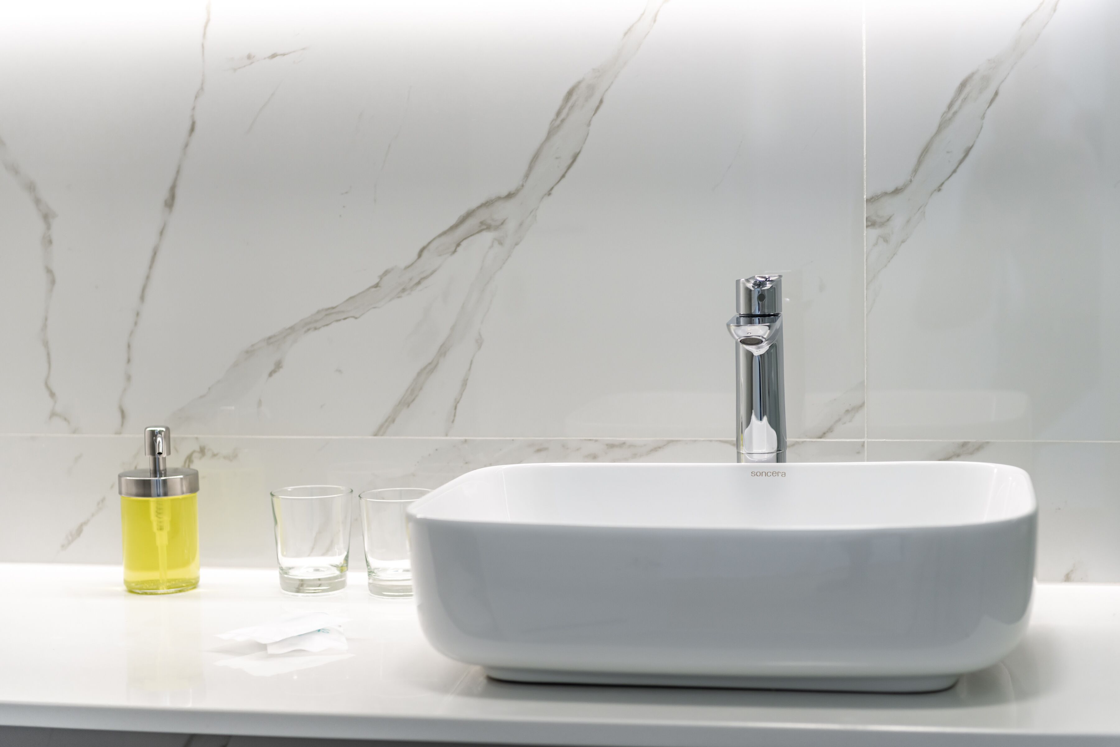 A bathroom with a white marble sink, a faucet, a bottle of liquid soap, and two glasses.