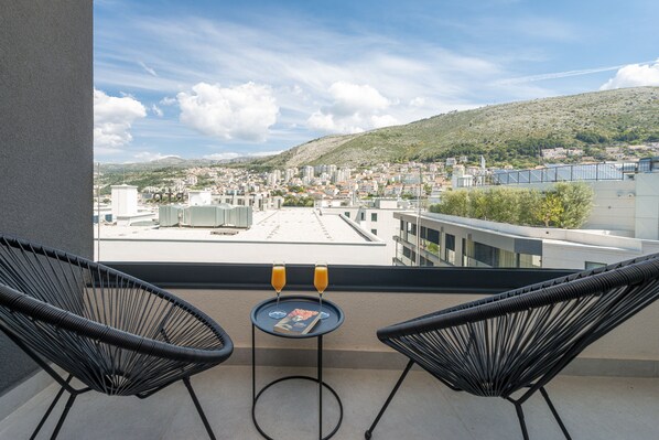 A balcony with two black wicker chairs, a small round table, and two glasses of orange juice. The view includes a cityscape with buildings and a hill in the distance. - City Residences (Dubrovnik)