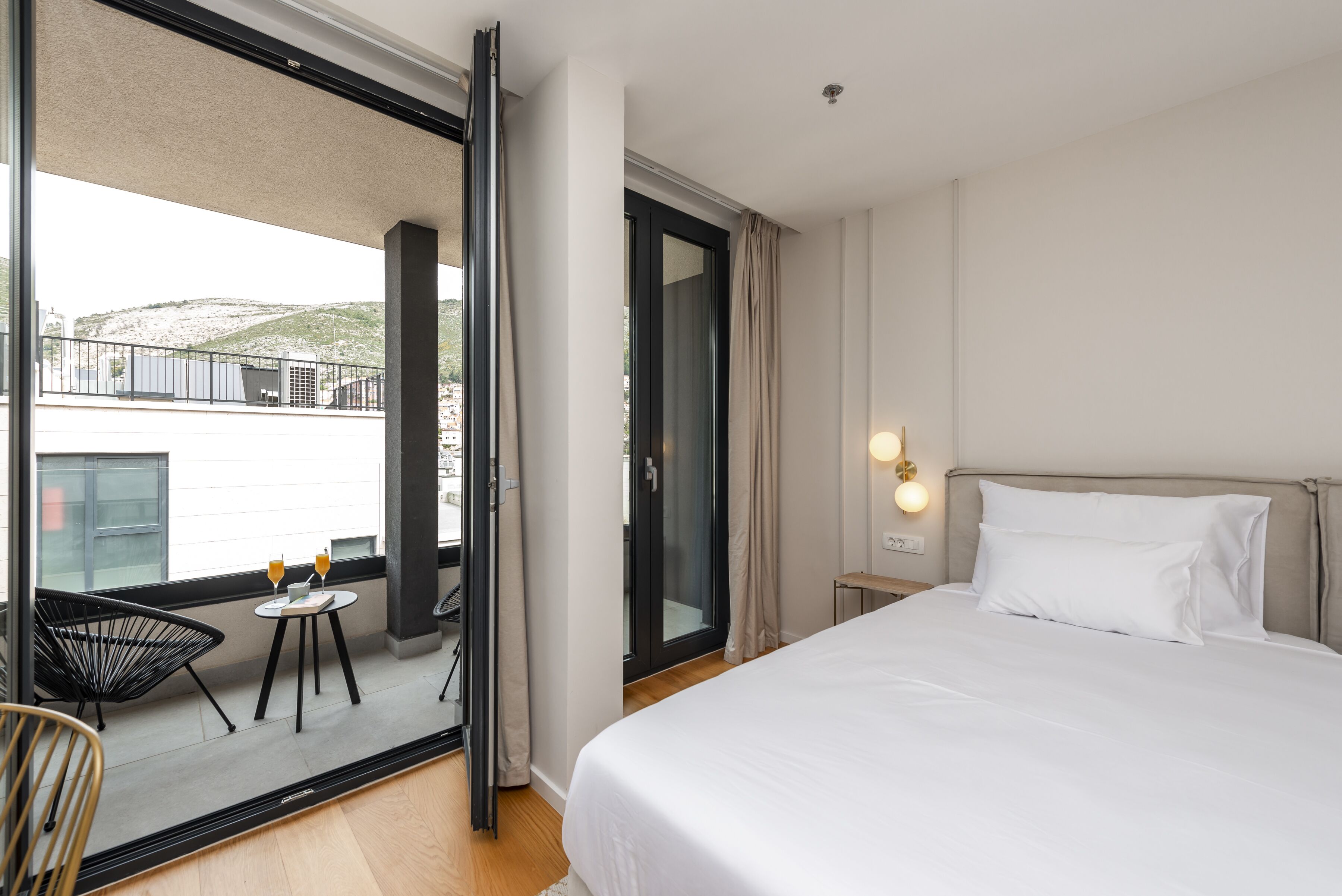 Une chambre d’hôtel avec un lit, une petite table sur laquelle sont posés deux verres de jus d’orange, une chaise et un balcon offrant une vue sur un immeuble et des collines.