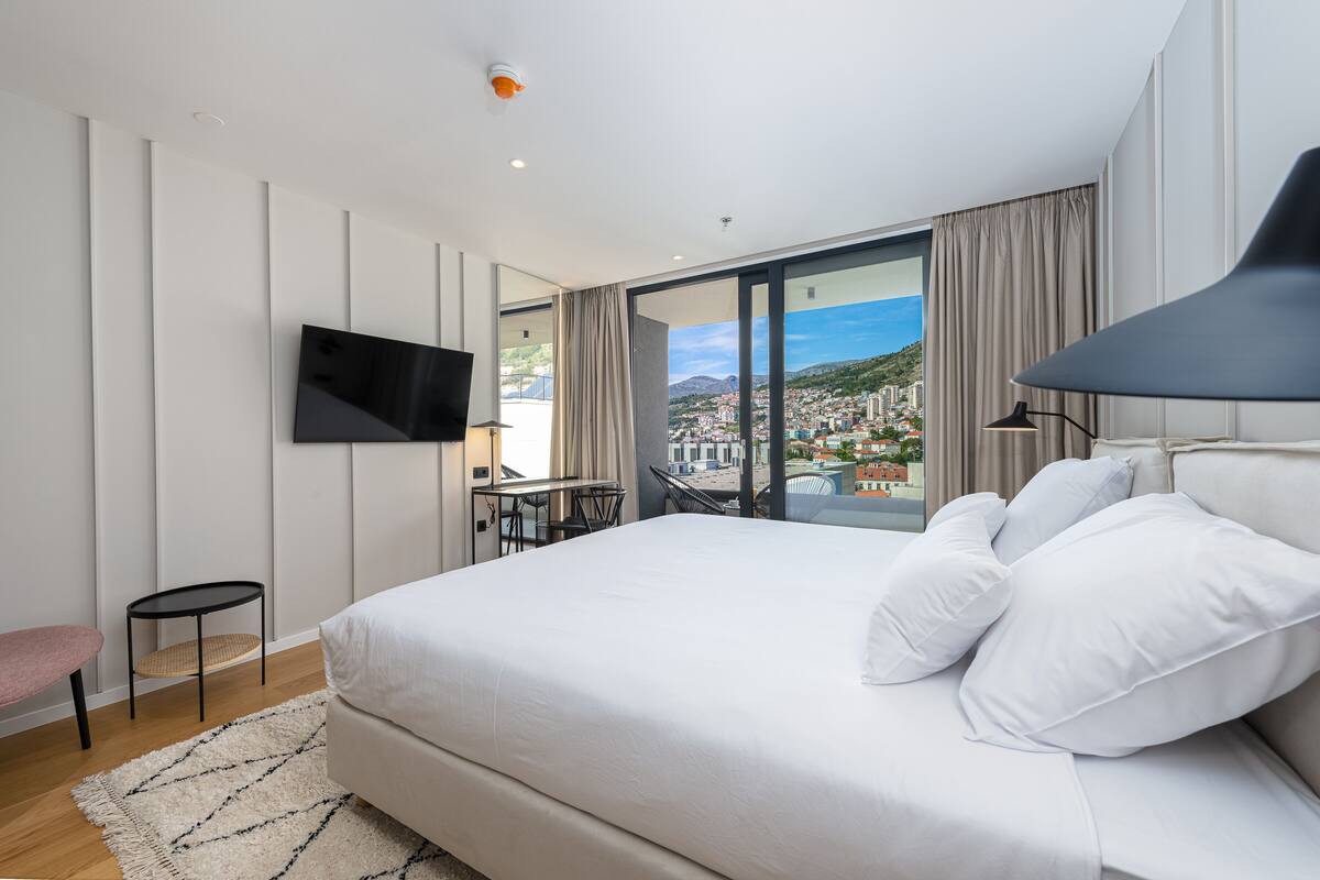 A modern hotel room with a large bed, a flat-screen TV mounted on the wall, a small table with chairs, and a view of a cityscape through large windows.