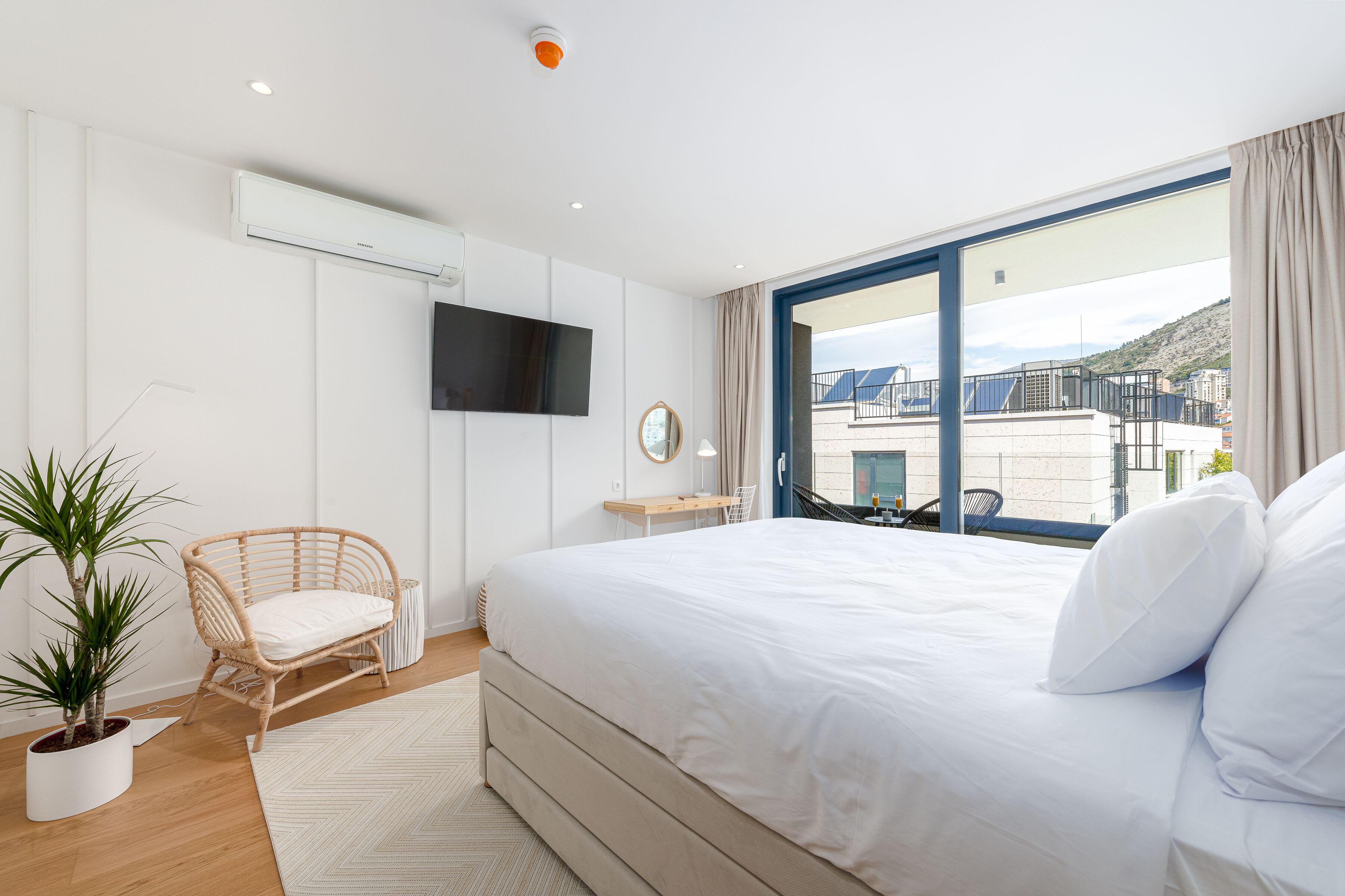 Un dormitorio moderno con una cama grande, un televisor de pantalla plana, una planta en maceta, una silla de mimbre y vistas de la ciudad a través de grandes ventanales.