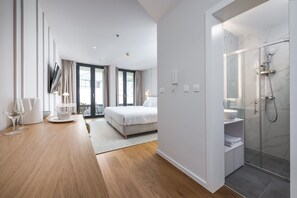 A modern hotel room with a large bed, a glass-enclosed shower, and a dining area with a table and chairs. - City Residences (Dubrovnik)