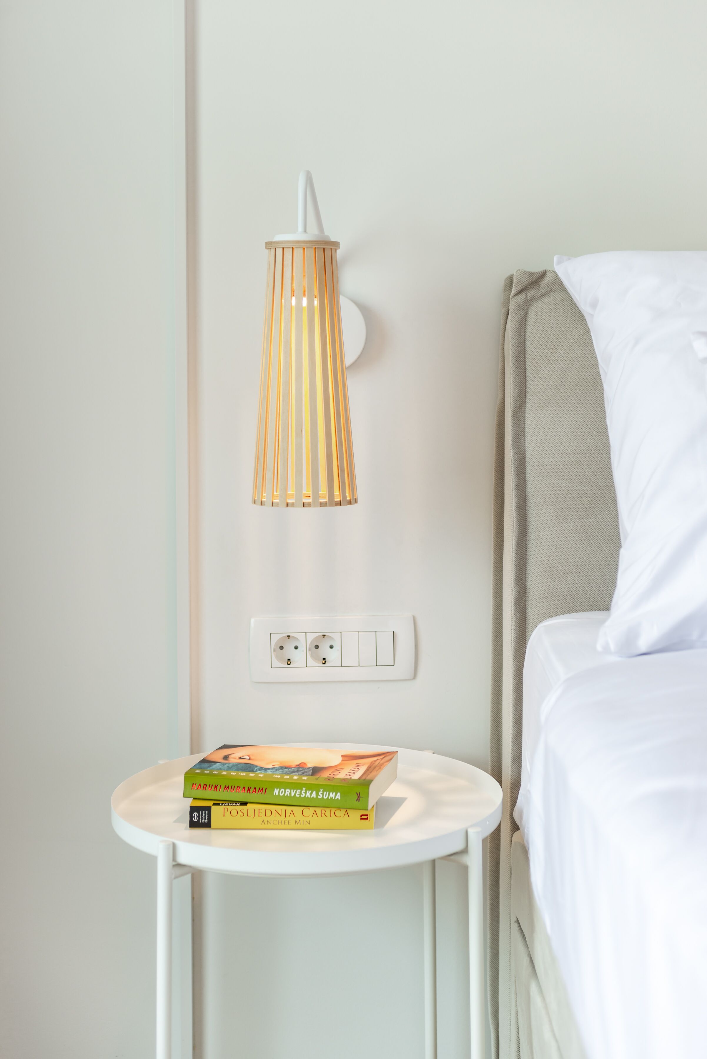 A neatly made bed with a white pillow and a beige headboard, a small bedside table with a lamp, and a power outlet.