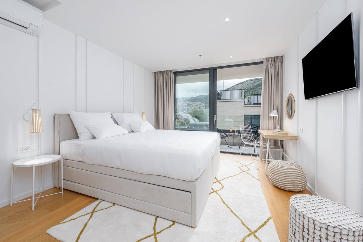 A modern bedroom with a large bed, a flat-screen TV, and a view of a cityscape through large windows.