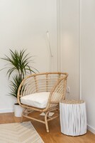 Uma cadeira de vime com almofada branca, uma planta em vaso e um banquinho tecido branco num quarto com soalho de madeira e paredes brancas.
