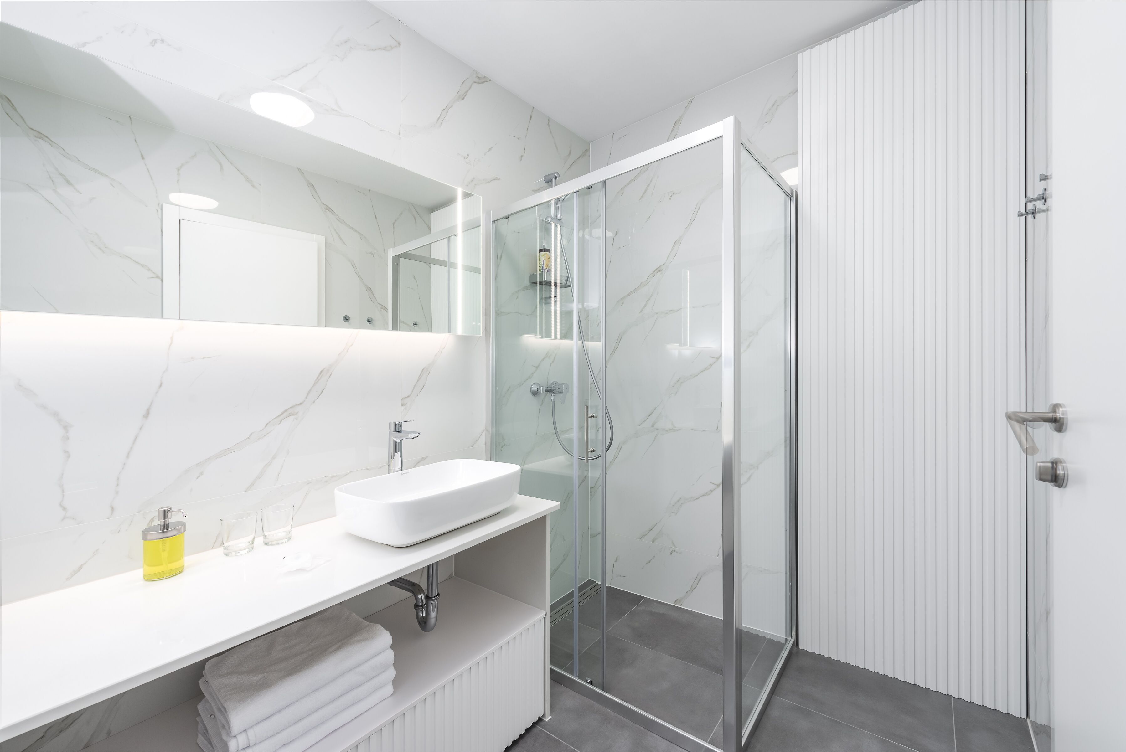 A modern bathroom with a white marble wall, a glass shower enclosure, a white sink, and a mirror above the sink.