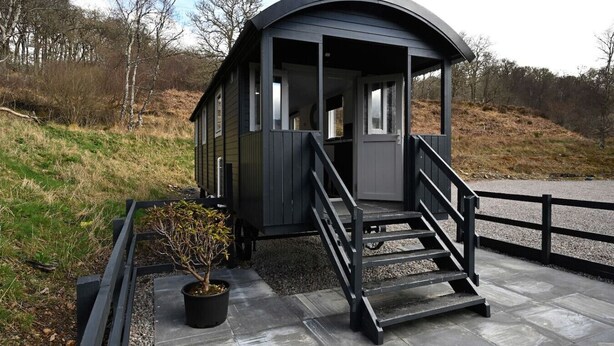 A small, dark-colored cabin with a curved roof and a set of stairs leading to a door. - The Carriage near Fort William (Fort William)