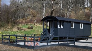 A small, dark-colored cabin with a covered porch, situated in a natural setting with trees and a grassy area. - The Carriage near Fort William (Fort William)
