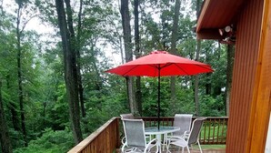 Une terrasse en bois avec une table et des chaises, placée sous un parasol rouge, et entourée d’une balustrade en bois et d’arbres.