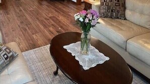 Un salon chaleureux avec des poutres en bois, un miroir rond, une commode, un canapé et une table basse agrémentée d’un vase de fleurs.