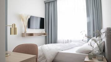 A modern hotel room with a bed, a desk, a TV, and a window with curtains.