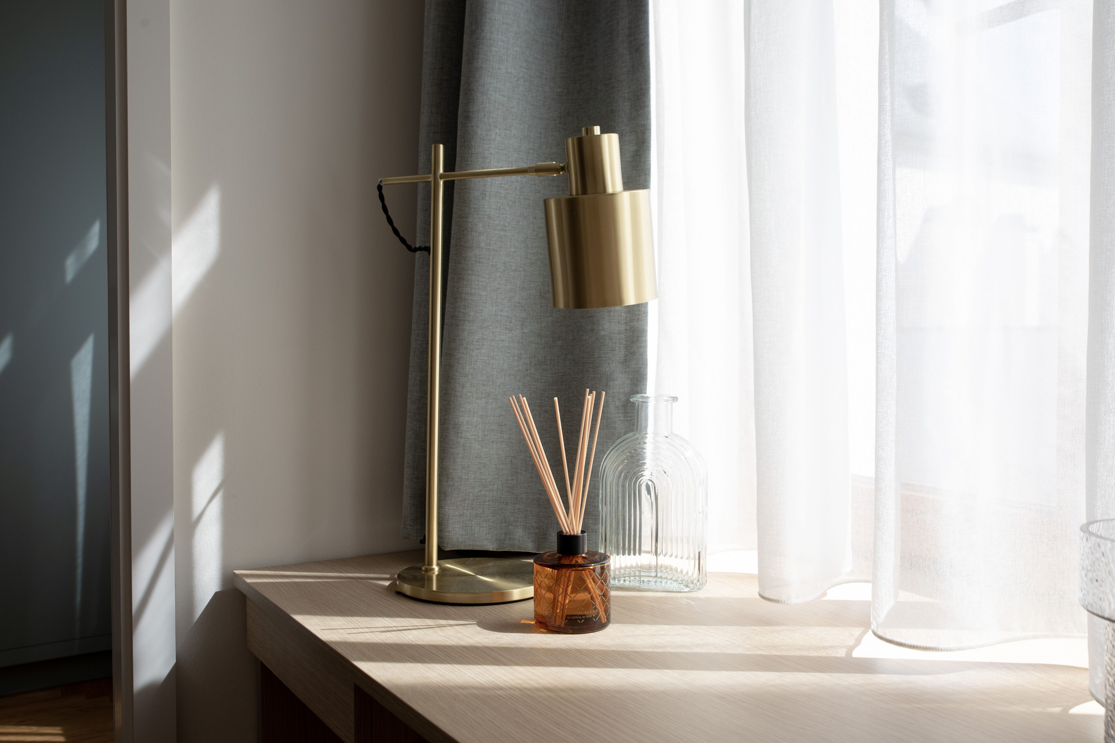 A wooden desk with a lamp, a glass bottle, and reed diffusers by a window with sheer curtains.
