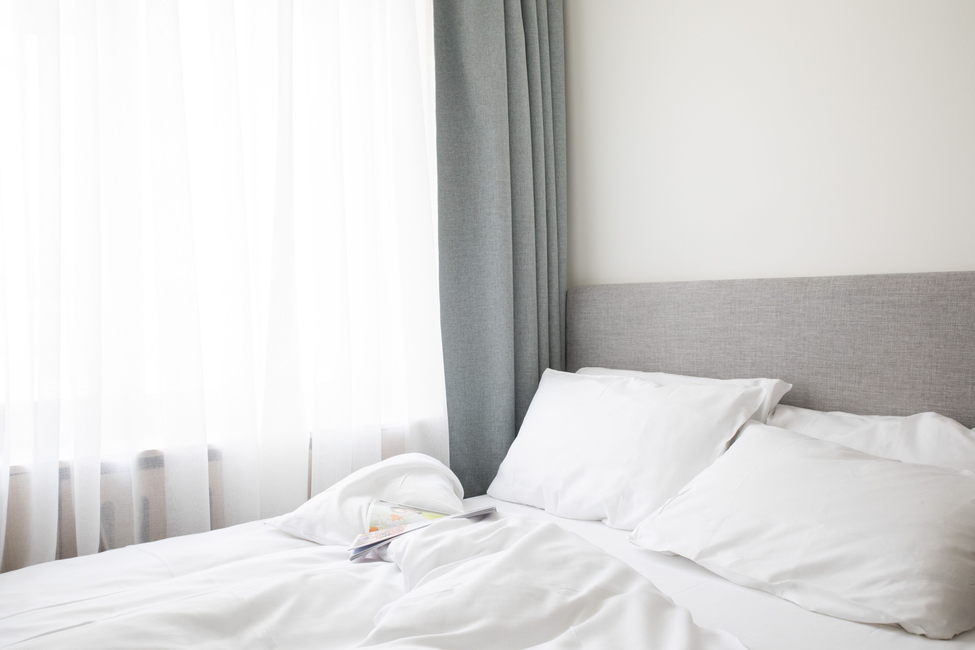 Ein ordentlich gemachtes Bett mit weißen Leinen und Kissen, ein grauer Kopfteil und leichte Vorhänge, die natürliches Licht hereinlassen.