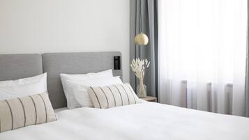 Ein ordentlich bezogenes Bett mit weißen Leinen und gestreiften Kissen, ein grauer Kopfteil und ein Nachttisch mit einer Lampe und einer Vase mit getrockneten Blumen.