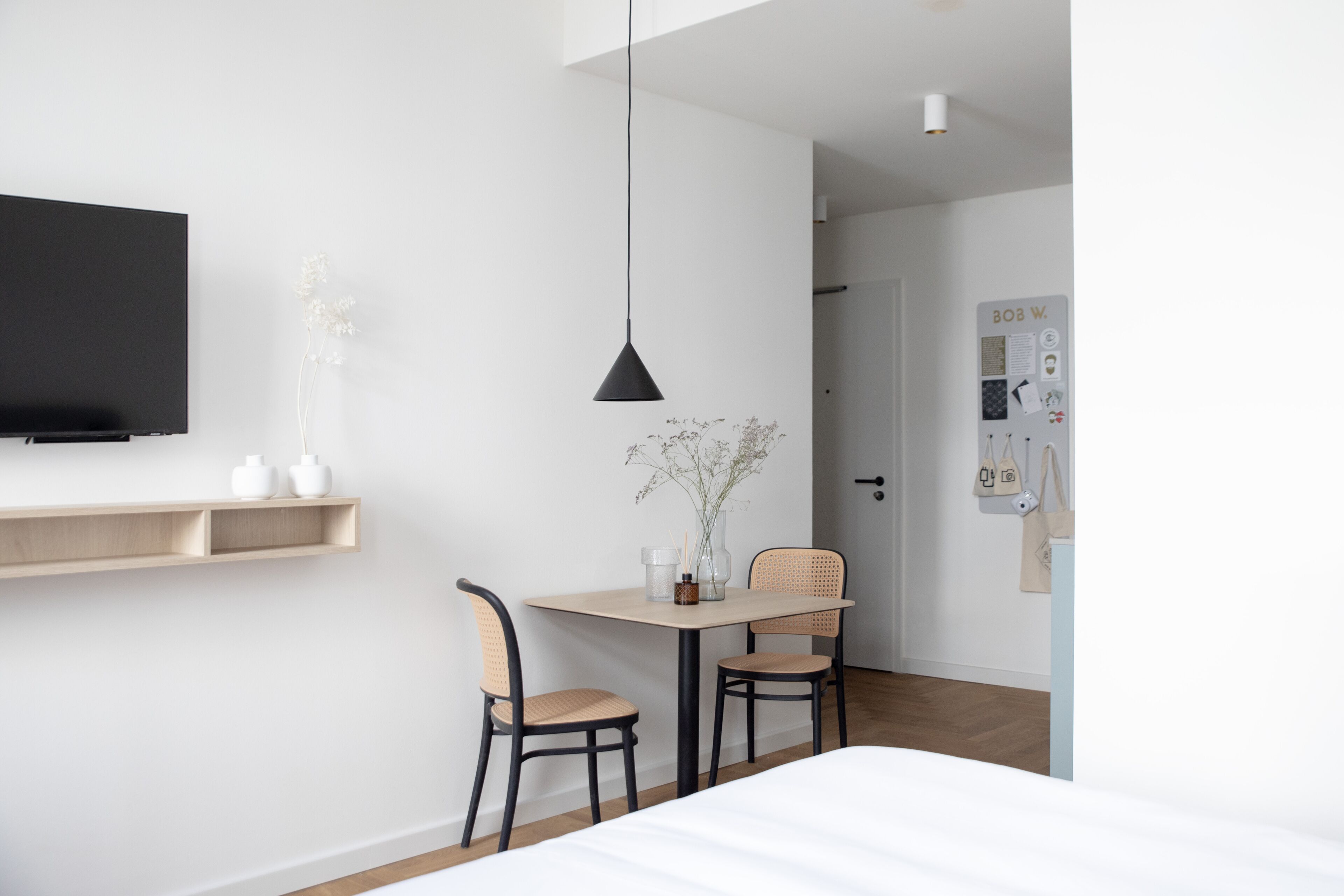 Una habitación de hotel moderna con una cama, un televisor instalado en la pared, una mesita con sillas y un tablón de anuncios en la pared.